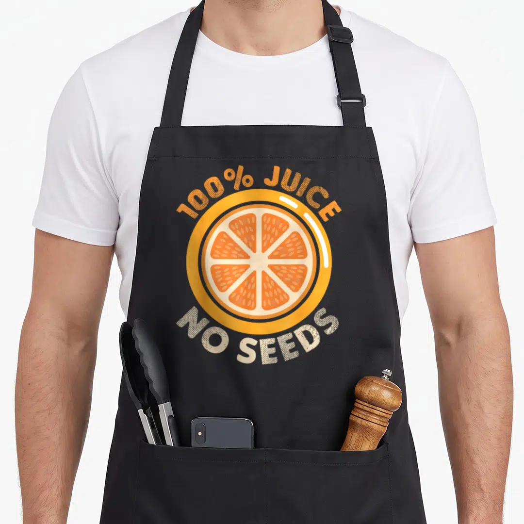 Vasectomy source apron - Cartoon orange slice with 100% JUICE NO SEEDS text in orange and off-white — shown on a black cotton canvas apron against a white background