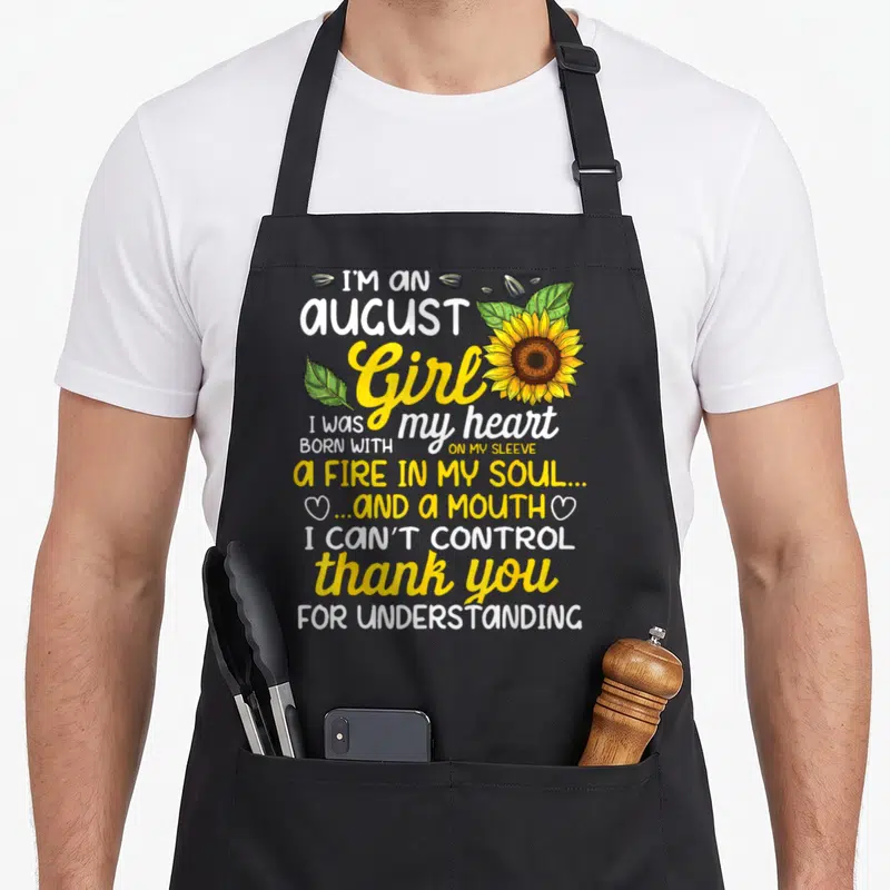 August sunflower apron - I'm an August Girl sunflower print with yellow white green text and scattered seeds — shown on a black cotton canvas apron against a white background