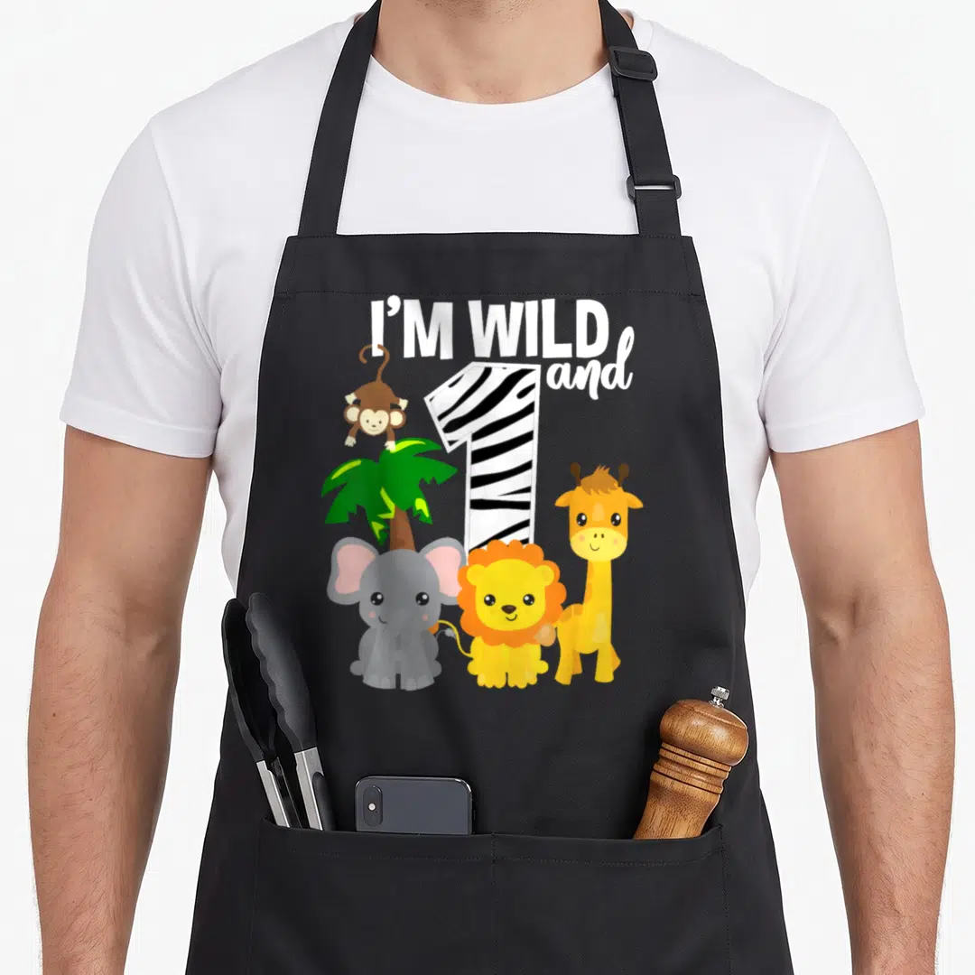 Wild zoo birthday apron - I'M WILD and 1 text with zebra stripe number 1, monkey, elephant, lion, giraffe, and palm — shown on a black cotton canvas apron against a white background