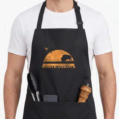 Dakota 70s apron - Orange retro sun with bison silhouette, three birds, and NORTH DAKOTA text — shown on a black cotton canvas apron against a white background