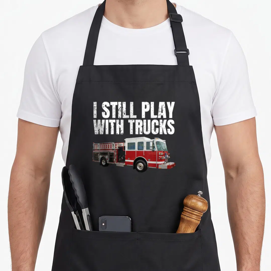 Play firefighter apron - I STILL PLAY WITH TRUCKS text above a red and white fire truck photo — shown on a black cotton canvas apron against a white background
