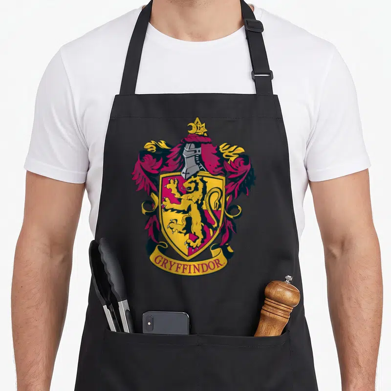 Funny crest novelty apron - Cartoon shield with lion, helmet, moon star, and GRYFFINDOR banner in red gold silver — shown on a black cotton canvas apron against a white background