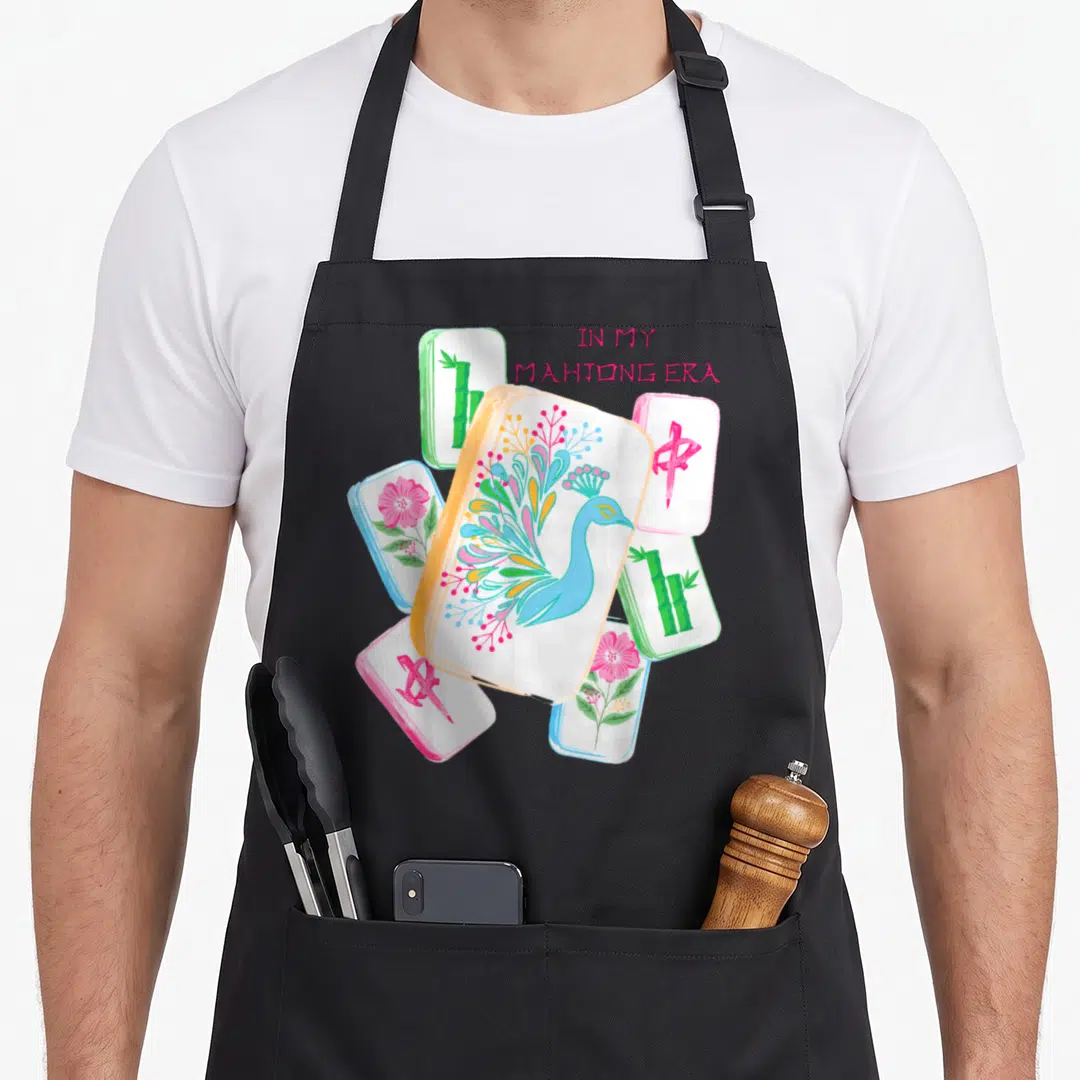 Era mahjong apron - Cartoon peacock and bamboo mahjong tiles with pink text IN MY MAHJONG ERA — shown on a black cotton canvas apron against a white background