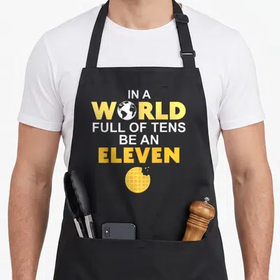 Ten eleven novelty apron - Yellow and white text reading IN A WORLD FULL OF TENS BE AN ELEVEN above a bitten waffle — shown on a black cotton canvas apron against a white background