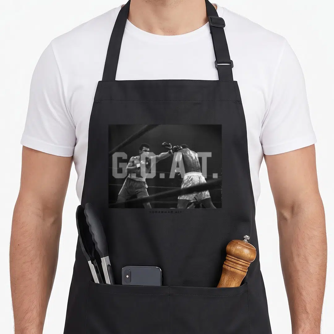 Muhammad goat apron - Black and white photo of two boxers with G.O.A.T. text overlay and Muhammad Ali caption below — shown on a black cotton canvas apron against a white background