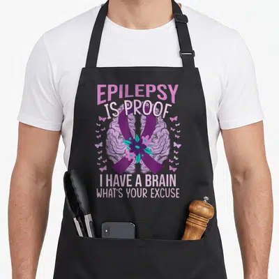 Vintage epilepsy awareness apron - Brain graphic with purple ribbon, teal flowers, butterflies, and text EPILEPSY IS — shown on a black cotton canvas apron against a white background