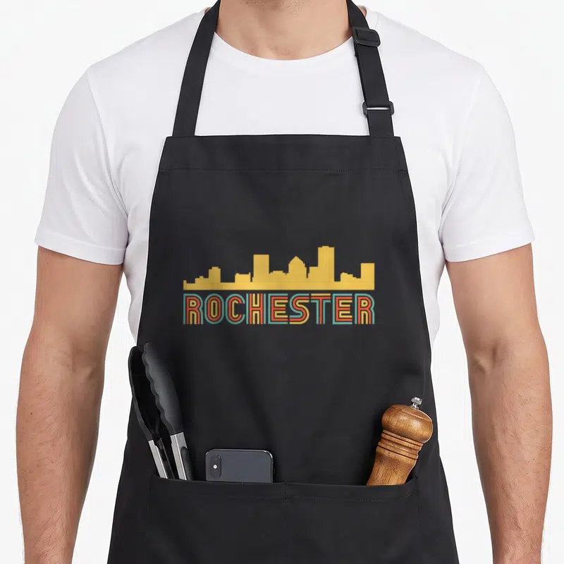 Rochester skyline new york apron - Yellow skyline silhouette above ROCHESTER text with red and teal outlines — shown on a black cotton canvas apron against a white background