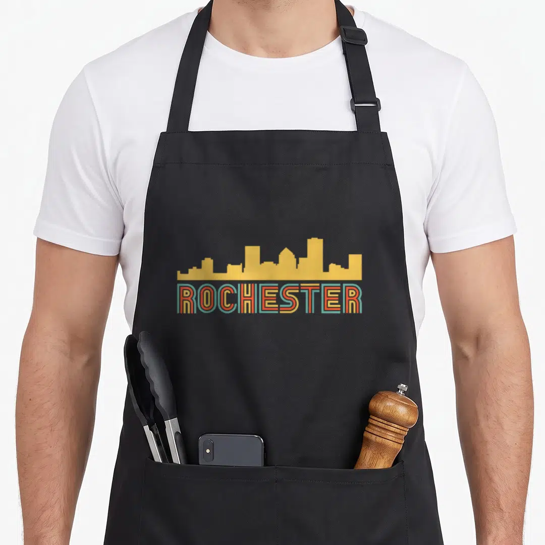 Rochester skyline new york apron - Yellow skyline silhouette above ROCHESTER text with red and teal outlines — shown on a black cotton canvas apron against a white background