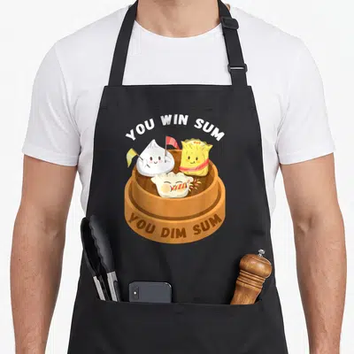 Win food aprons - Three cartoon dumplings in a brown steamer with You Win Sum You Dim Sum text — shown on a black cotton canvas apron against a white background