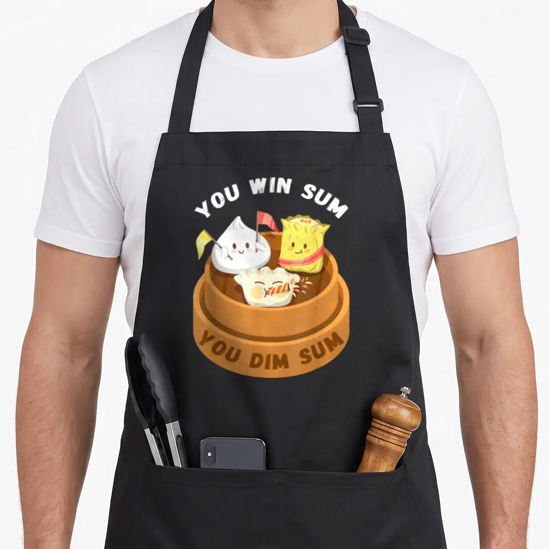 Win food aprons - Three cartoon dumplings in a brown steamer with You Win Sum You Dim Sum text — shown on a black cotton canvas apron against a white background