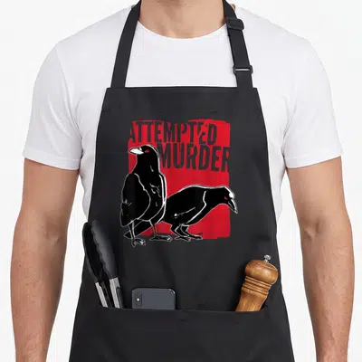 Pun ravens apron - Two black crows on red background with grey Attempted Murder text — shown on a black cotton canvas apron against a white background