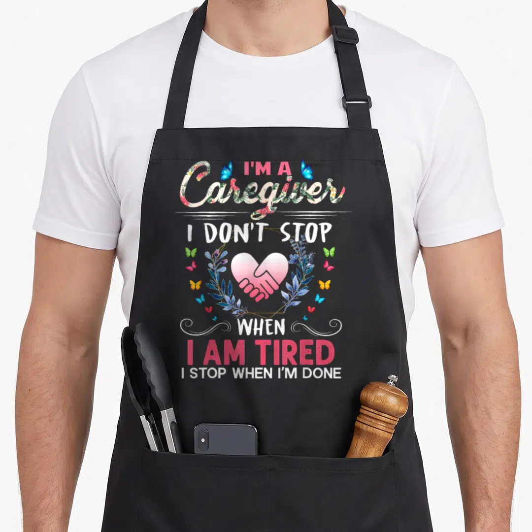Stop caregiver apron - Vertical text I'm a Caregiver I Don't Stop When I Am Tired with pink heart and blue leaves — shown on a black cotton canvas apron against a white background