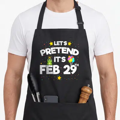 Feb 29th birthday apron - Cartoon frog with crown and balloons next to white text LET'S PRETEND IT'S FEB 29TH — shown on a black cotton canvas apron against a white background