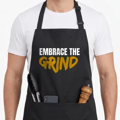 Embrace grindset apron - Stacked white and gold text reading EMBRACE THE GRIND — shown on a black cotton canvas apron against a white background