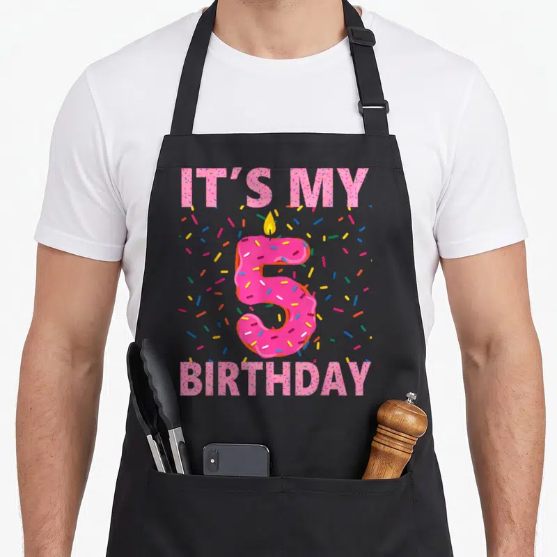 5th donut apron - Pink frosted number 5 with candle surrounded by IT'S MY BIRTHDAY text and colorful sprinkles — shown on a black cotton canvas apron against a white background
