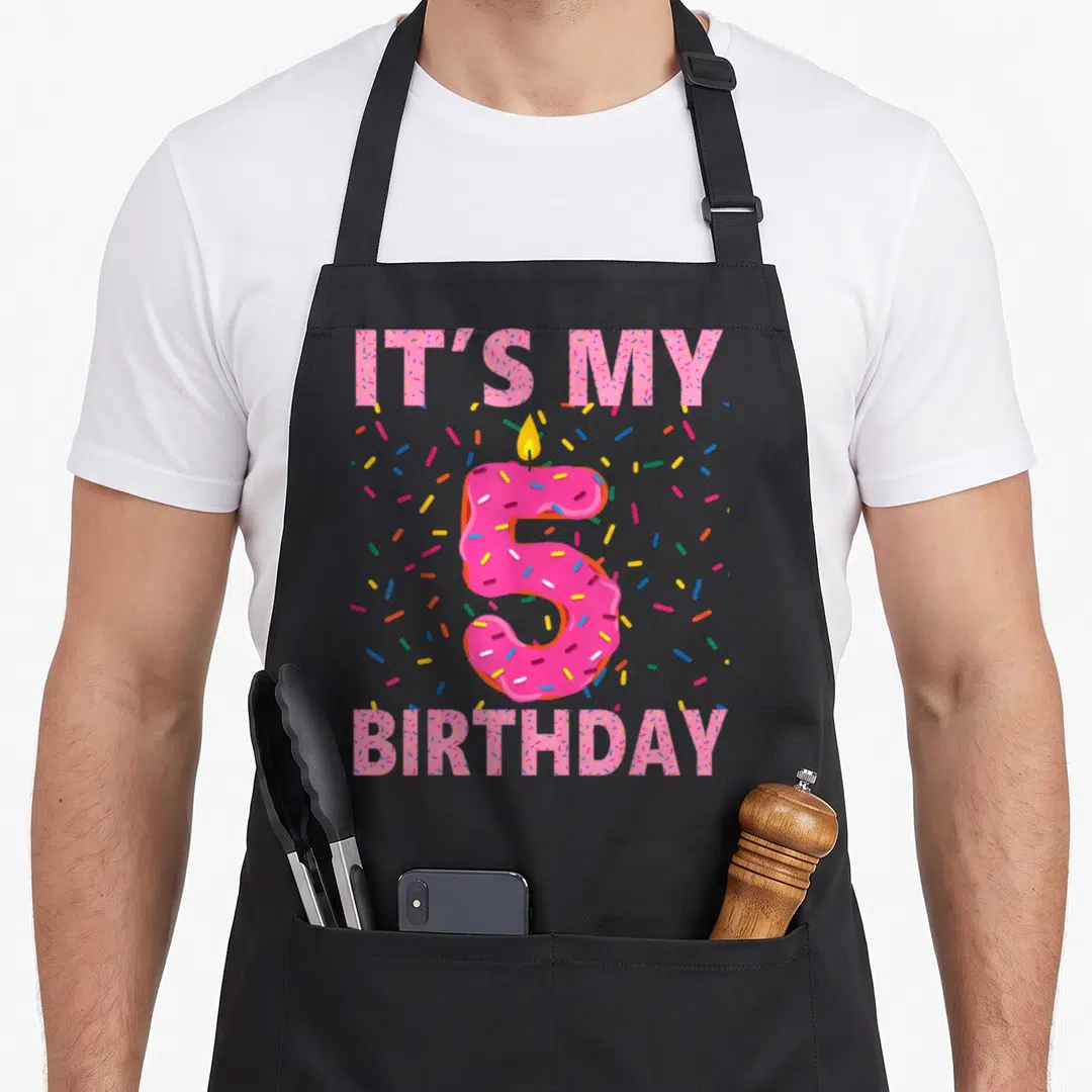 5th donut apron - Pink frosted number 5 with candle surrounded by IT'S MY BIRTHDAY text and colorful sprinkles — shown on a black cotton canvas apron against a white background