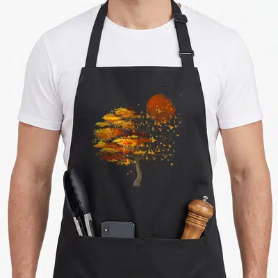 Flock tree apron - Watercolor tree with orange sun and scattered birds in autumn colors — shown on a black cotton canvas apron against a white background