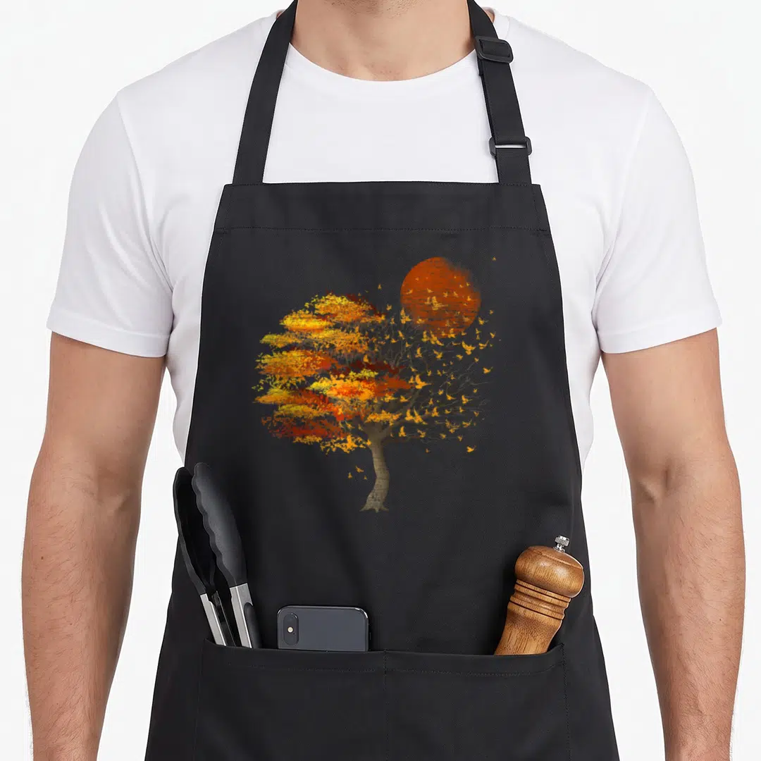 Flock tree apron - Watercolor tree with orange sun and scattered birds in autumn colors — shown on a black cotton canvas apron against a white background