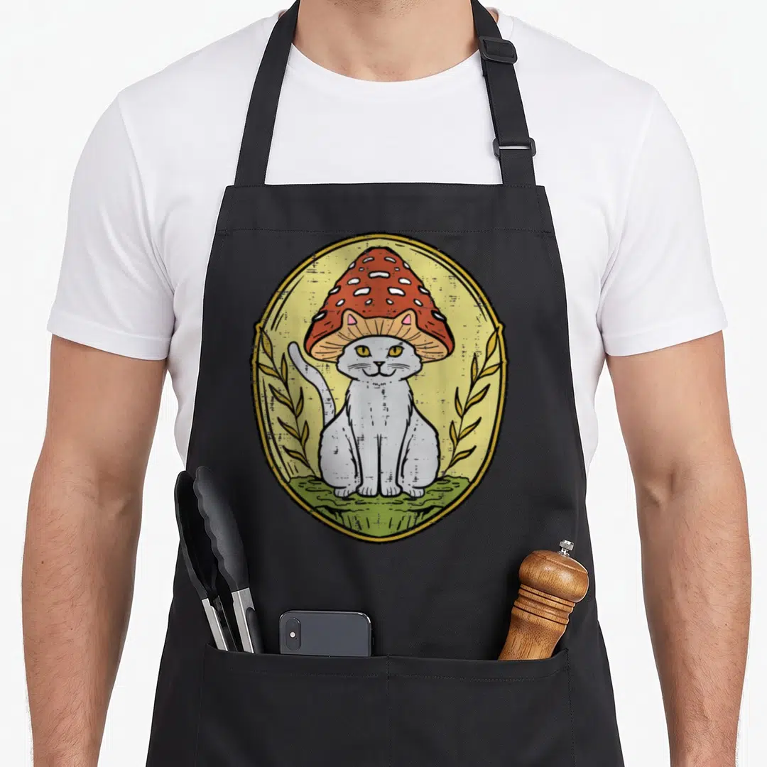 Funny cat cottagecore apron - Cartoon white cat wearing red spotted mushroom hat inside yellow oval frame with two — shown on a black cotton canvas apron against a white background