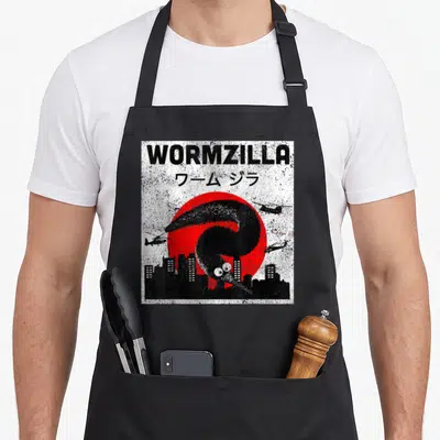 Worm magic apron - WORMZILLA text above giant black worm and red sun with helicopters over city — shown on a black cotton canvas apron against a white background