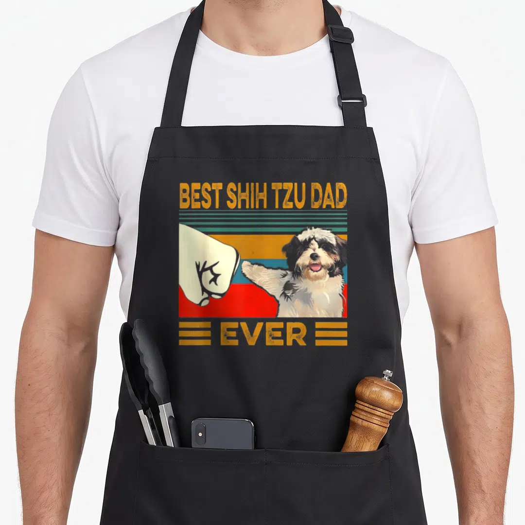 Shih dad mode apron - Retro graphic of human fist bumping a Shih Tzu paw with yellow and red stripes — shown on a black cotton canvas apron against a white background