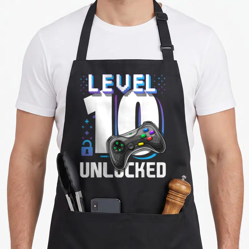 Video gamer 10th birthday apron - LEVEL 10 UNLOCKED text with game controller and blue lock icon — shown on a black cotton canvas apron against a white background