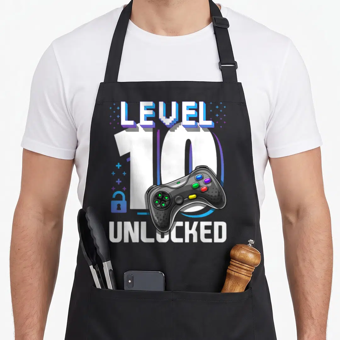 Video gamer 10th birthday apron - LEVEL 10 UNLOCKED text with game controller and blue lock icon — shown on a black cotton canvas apron against a white background