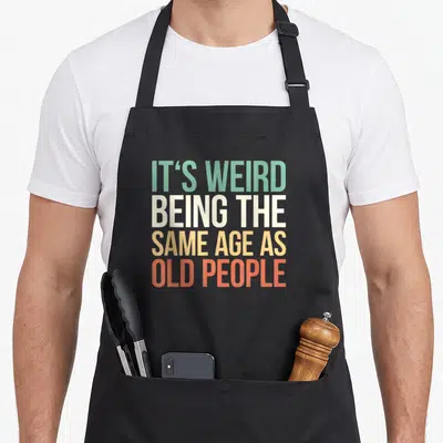 Retro age general weird aprons - Stacked text reading IT'S WEIRD BEING THE SAME AGE AS OLD PEOPLE in green white yellow — shown on a black cotton canvas apron against a white background