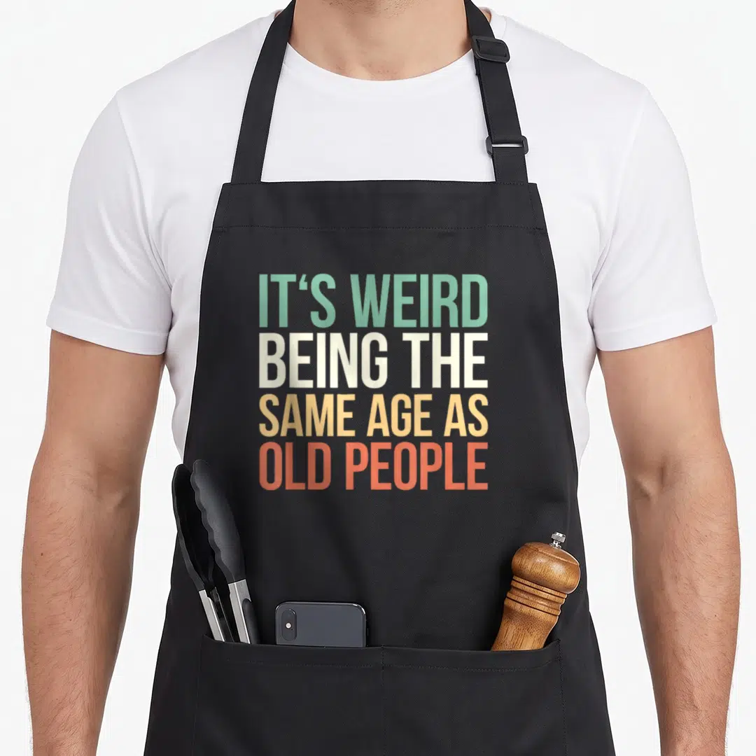 Retro age general weird aprons - Stacked text reading IT'S WEIRD BEING THE SAME AGE AS OLD PEOPLE in green white yellow — shown on a black cotton canvas apron against a white background