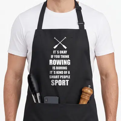Kayak crew apron - Two crossed white oars above seven lines of text reading IT'S OKAY IF YOU THINK ROWING IS BORING — shown on a black cotton canvas apron against a white background