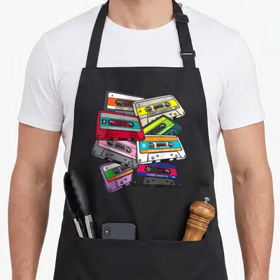 Generation 1980s apron - Seven overlapping cartoon cassette tapes in red green purple beige and black with stereo labels — shown on a black cotton canvas apron against a white background