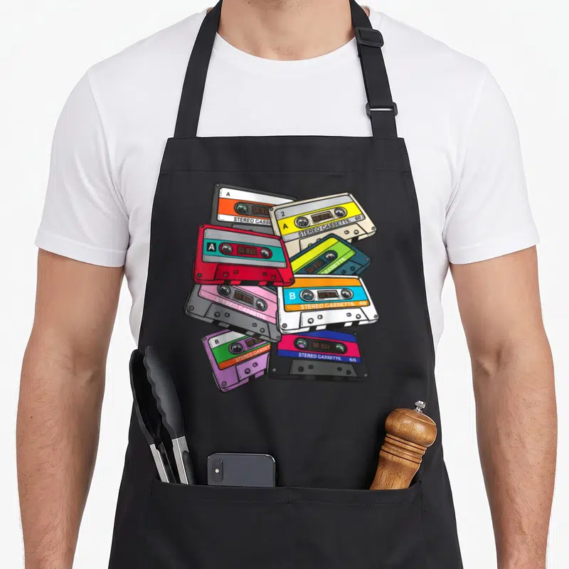 Generation 1980s apron - Seven overlapping cartoon cassette tapes in red green purple beige and black with stereo labels — shown on a black cotton canvas apron against a white background