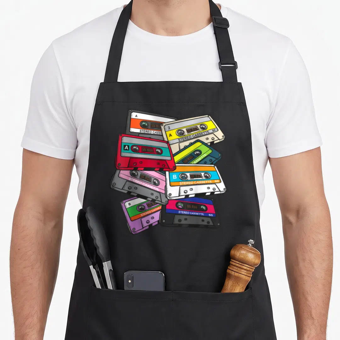 Generation 1980s apron - Seven overlapping cartoon cassette tapes in red green purple beige and black with stereo labels — shown on a black cotton canvas apron against a white background