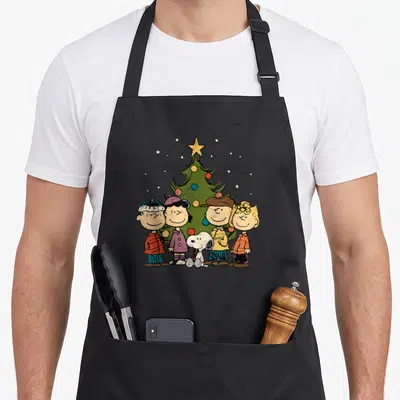 Funny christmas apron - Cartoon four children and dog standing in front of green Christmas tree with star and colored — shown on a black cotton canvas apron against a white background