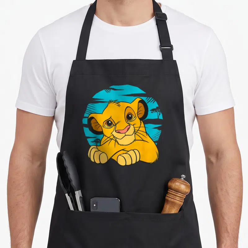 Young 90s apron - Yellow cartoon lion cub resting paws on blue textured circle background — shown on a black cotton canvas apron against a white background