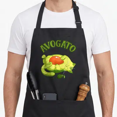 Meow mexico apron - Arched AVOGATO text over light green cat lying on avocado with orange pit and green yarn ball — shown on a black cotton canvas apron against a white background