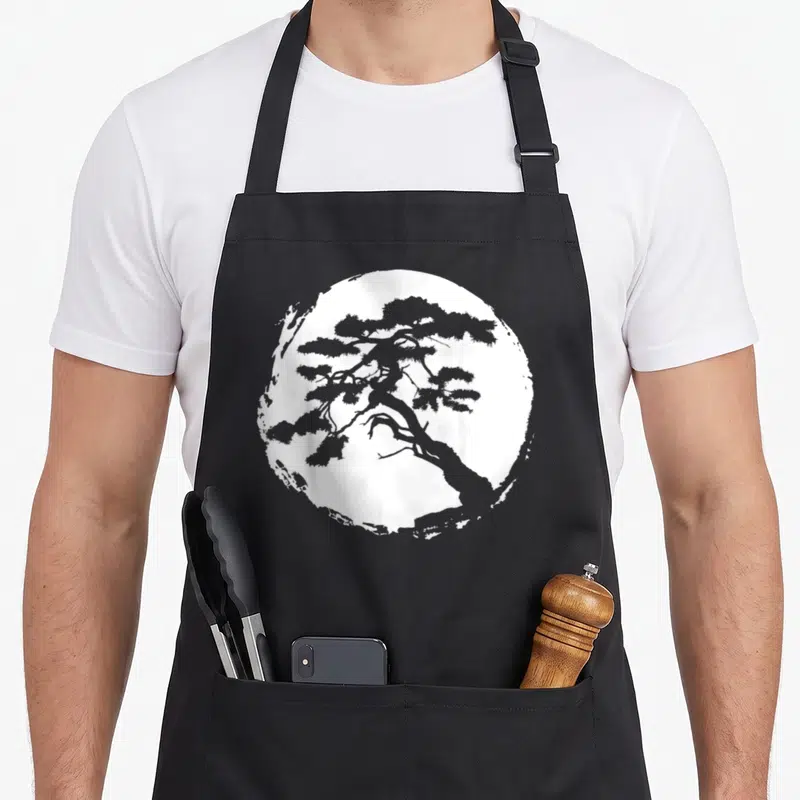 Bonsai master apron - Grey pine tree silhouette inside a distressed white circle on black fabric — shown on a black cotton canvas apron against a white background
