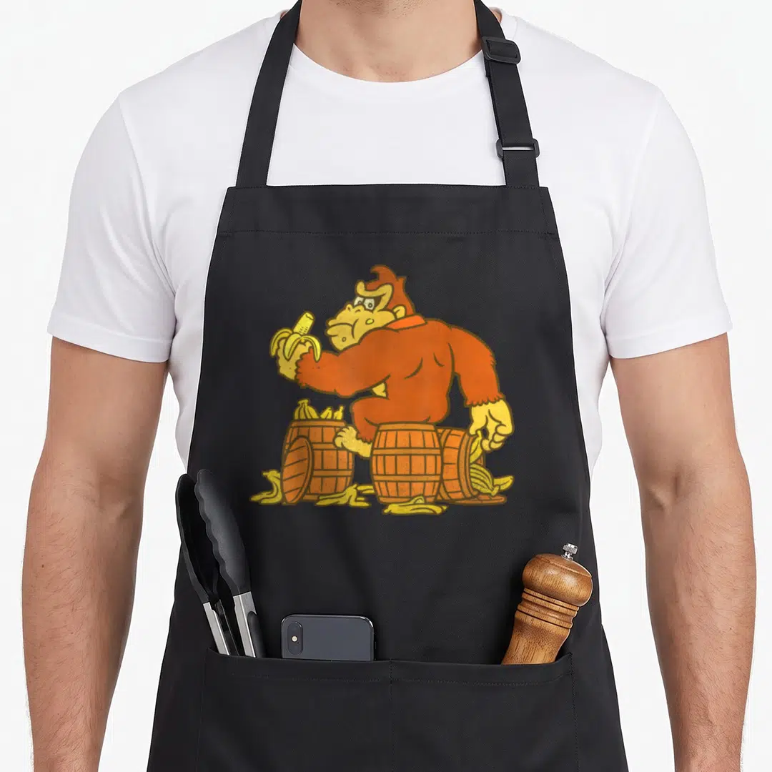 Bananas barrel apron - Cartoon gorilla in orange sitting on three wooden barrels holding yellow bananas — shown on a black cotton canvas apron against a white background