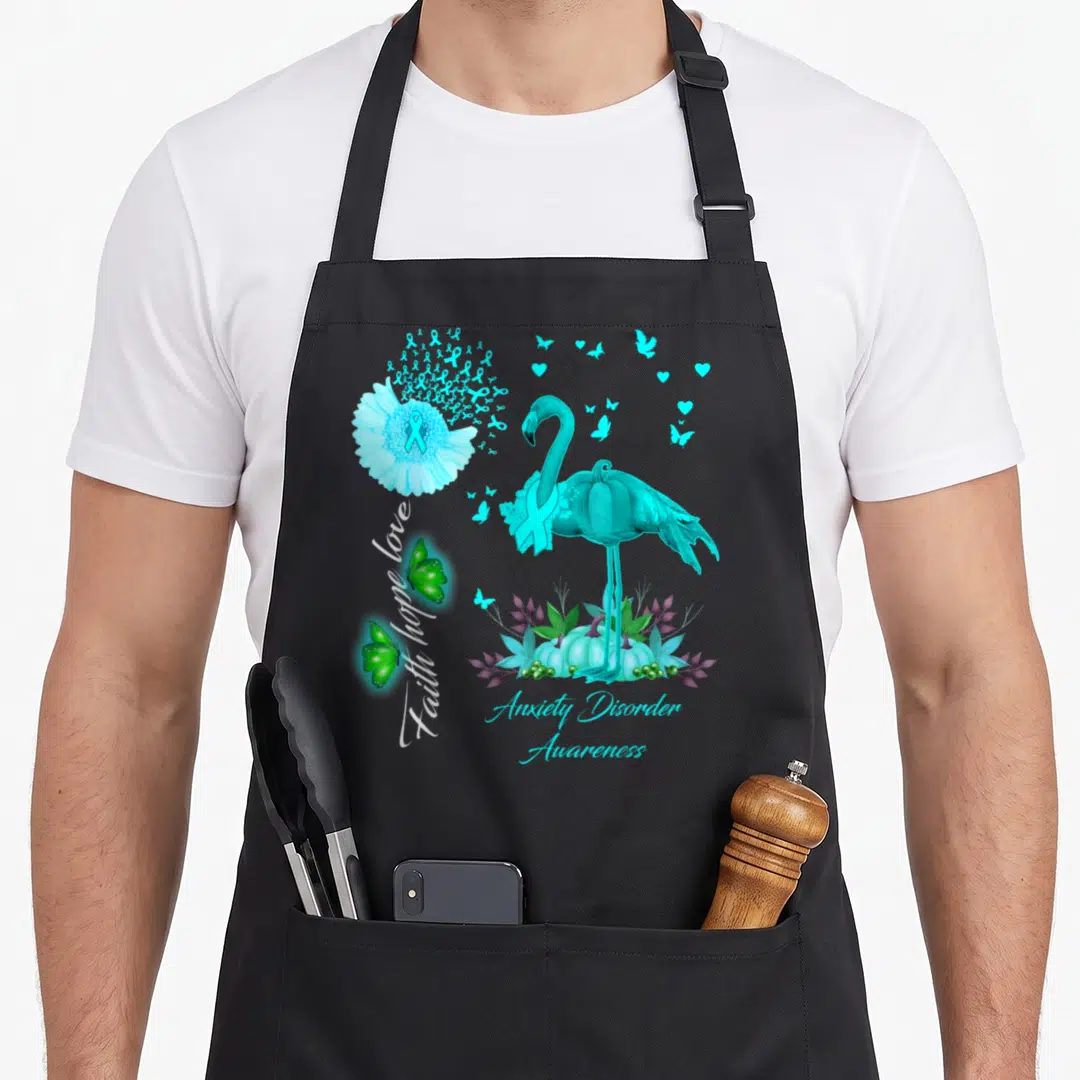Faith flamingo apron - Cyan flamingo with ribbon, green butterflies, purple leaves, and anxiety disorder awareness text — shown on a black cotton canvas apron against a white background