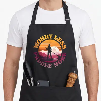 Worry paddleboard apron - Retro sunset circle with female paddleboarder silhouette and arching WORRY LESS PADDLE MORE — shown on a black cotton canvas apron against a white background
