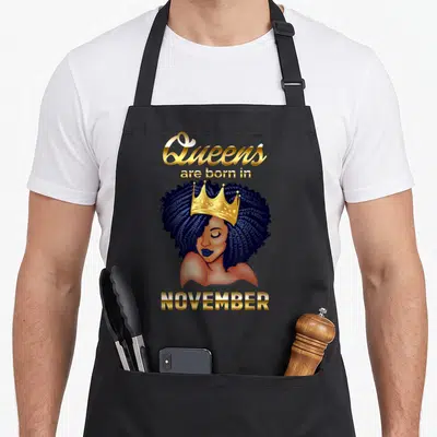 Queen november birthday apron - Crowned Black woman with blue hair and gold text Queens are born in NOVEMBER — shown on a black cotton canvas apron against a white background