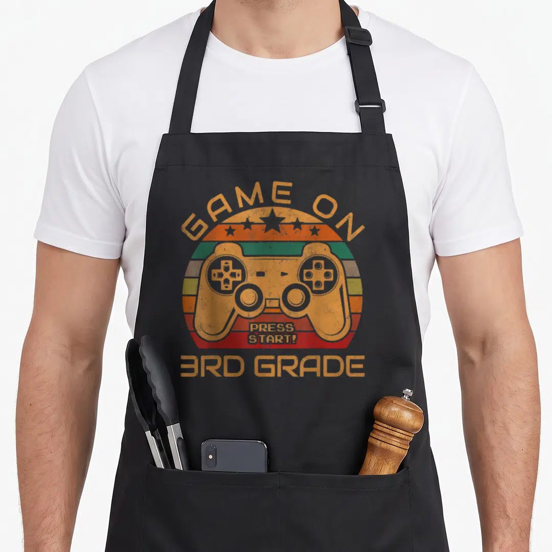 3rd game aprons - Retro orange controller over striped semi-circle with GAME ON PRESS START 3RD GRADE text — shown on a black cotton canvas apron against a white background