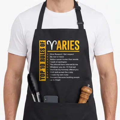 Birthday aries kitchen apron - Yellow and white typography listing ten Aries rules including sarcasm and honesty — shown on a black cotton canvas apron against a white background