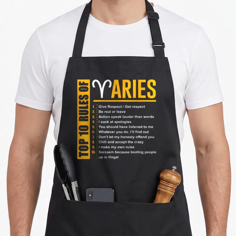 Birthday aries kitchen apron - Yellow and white typography listing ten Aries rules including sarcasm and honesty — shown on a black cotton canvas apron against a white background