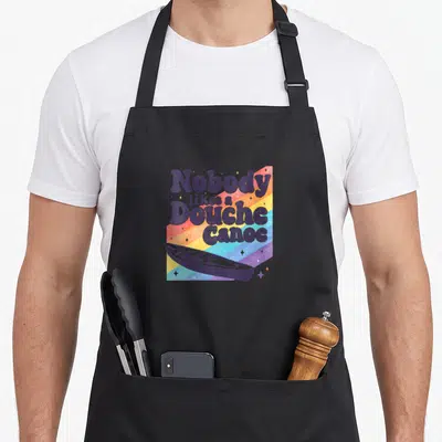 Douche vacation beach apron - Retro purple text reading Nobody likes a Douche Canoe over rainbow stripes and boat — shown on a black cotton canvas apron against a white background