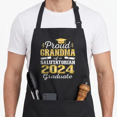 Salutatorian grandma apron - Gold graduation cap and two white diplomas flanking proud grandma salutatorian 2024 text — shown on a black cotton canvas apron against a white background