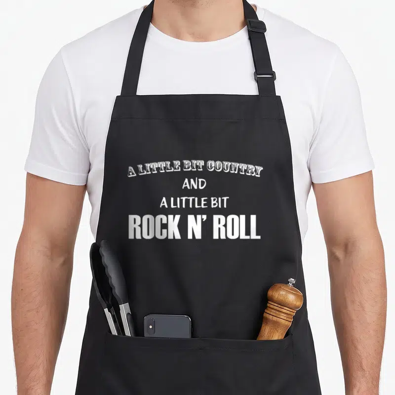 Bit country apron - White stacked text reading A Little Bit Country and a Little Bit Rock N' Roll — shown on a black cotton canvas apron against a white background