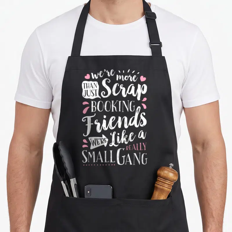 Scrapbooking friends apron - Stacked white and pink text reading we're more than just scrap booking friends surrounded — shown on a black cotton canvas apron against a white background