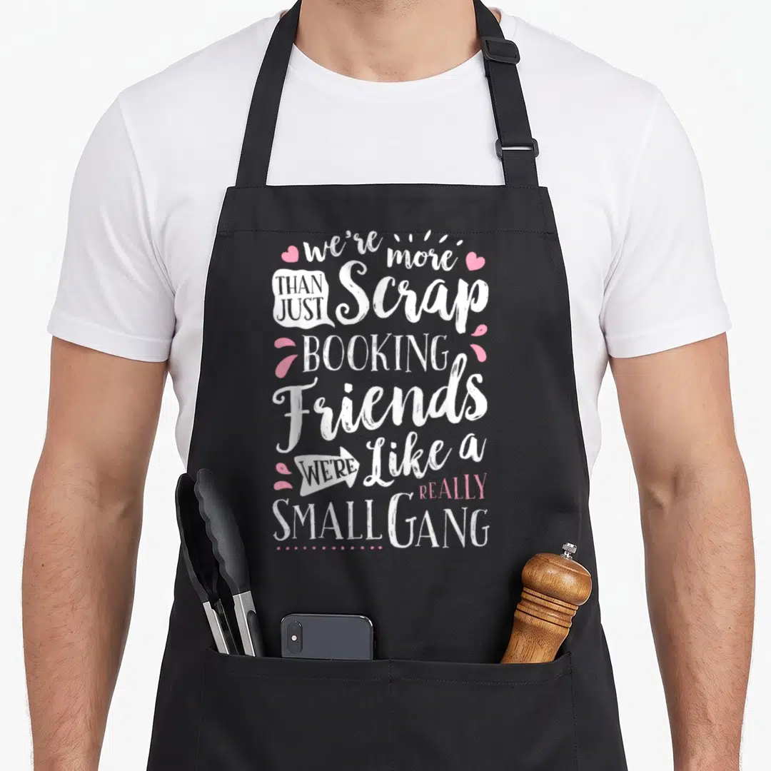 Scrapbooking friends apron - Stacked white and pink text reading we're more than just scrap booking friends surrounded — shown on a black cotton canvas apron against a white background