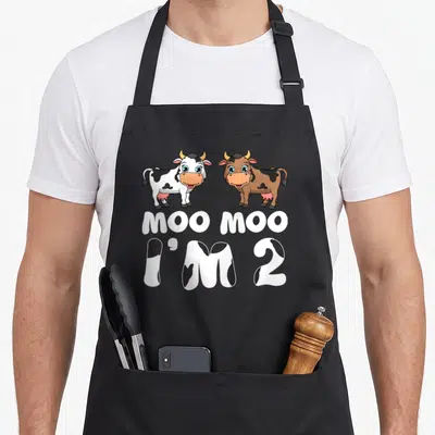 2nd cow apron - Two cartoon cows above MOO MOO I'M 2 text with cow spot pattern fill — shown on a black cotton canvas apron against a white background